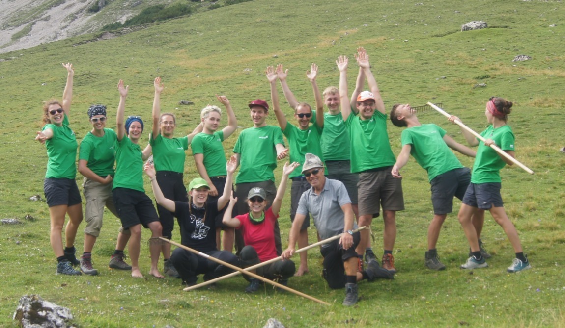 Eine Gruppe von Menschen in grünen Hemden posiert auf einem grasbewachsenen Hügel bei den Zirler Almen und reckt fröhlich die Arme in die Höhe, während zwei Personen mit Holzstöcken davor knien.