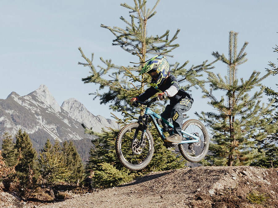 Eine Person, die einen Helm und eine Ausrüstung trägt, fährt mit einem Mountainbike eine aufregende Downhill-Strecke in einem Bikepark hinunter, umgeben von immergrünen Bäumen und Bergen im Hintergrund.