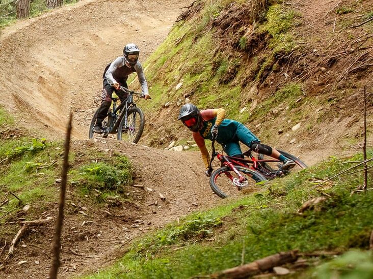 Zwei behelmte Mountainbiker fahren auf einem unbefestigten Weg durch einen bewaldeten Bikepark, wobei sie geschickt einen kurvigen Abschnitt des Weges bewältigen.