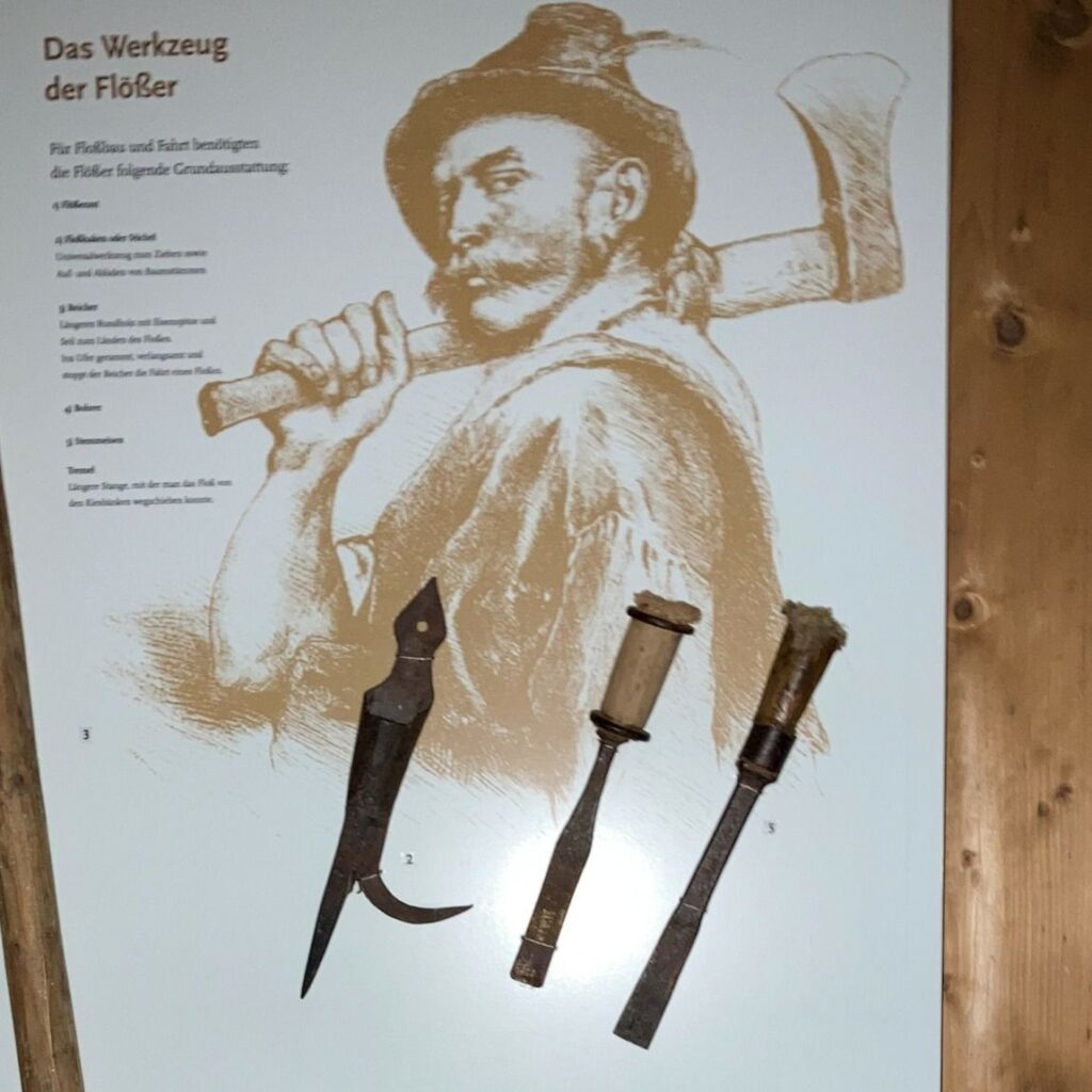 Schautafel mit "Das Werkzeug der Flößer": eine Zeichnung eines Mannes mit einer Axt und drei originale Flößerwerkzeuge mit den Bezeichnungen 1, 2 und 3, die auf der Tafel angebracht und beschriftet sind.