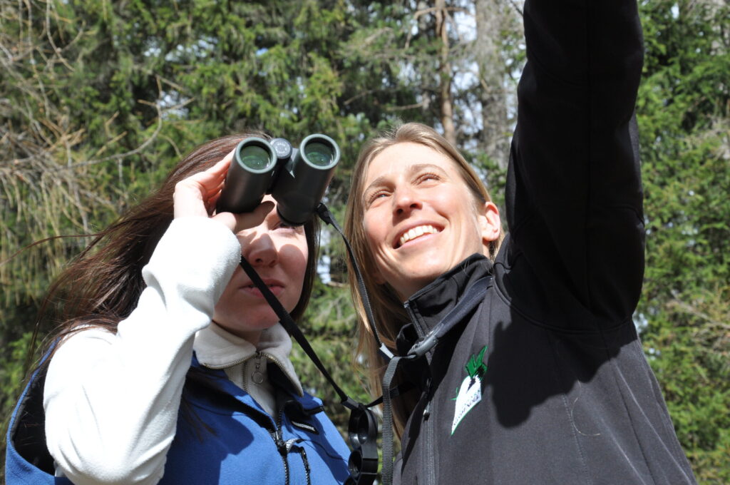 Zwei Frauen sind im Freien; eine benutzt ein Fernglas, während die andere nach oben gestikuliert. Beide scheinen mit der Neugier eines Naturpark-Rangers etwas in den Bäumen zu beobachten.