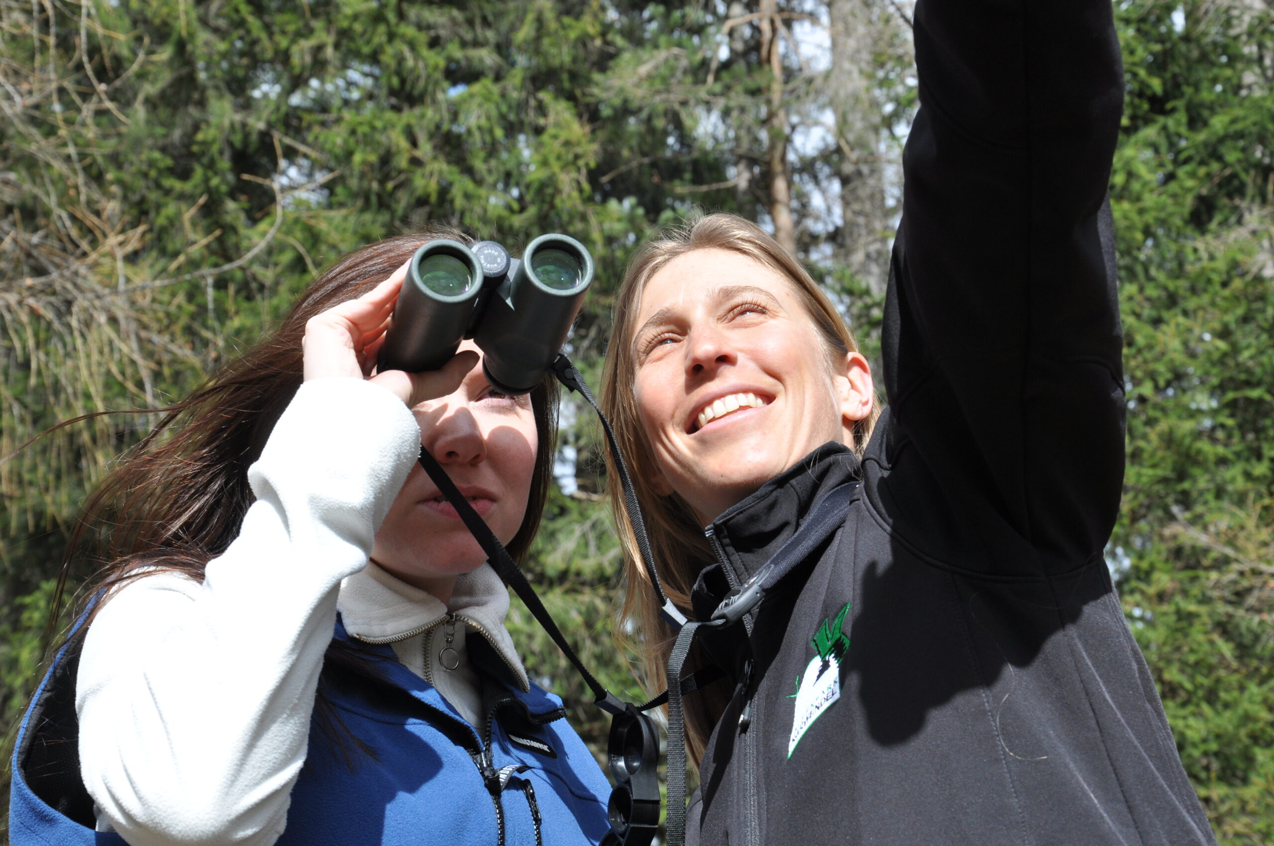 Zwei Frauen sind im Freien; eine benutzt ein Fernglas, während die andere nach oben gestikuliert. Beide scheinen mit der Neugier eines Naturpark-Rangers etwas in den Bäumen zu beobachten.