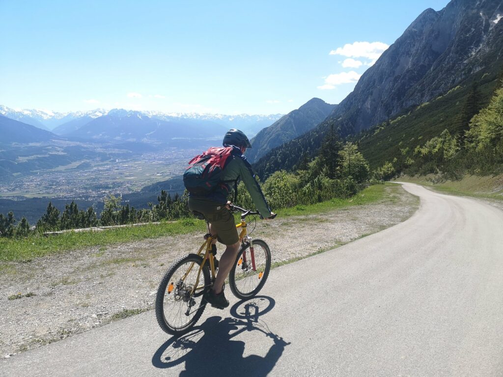 Eine Person, die einen Helm und einen Rucksack trägt, fährt mit dem Fahrrad auf einer Bergstraße in der Nähe der Hinterhornalm, mit einem malerischen Tal und Bergen im Hintergrund.