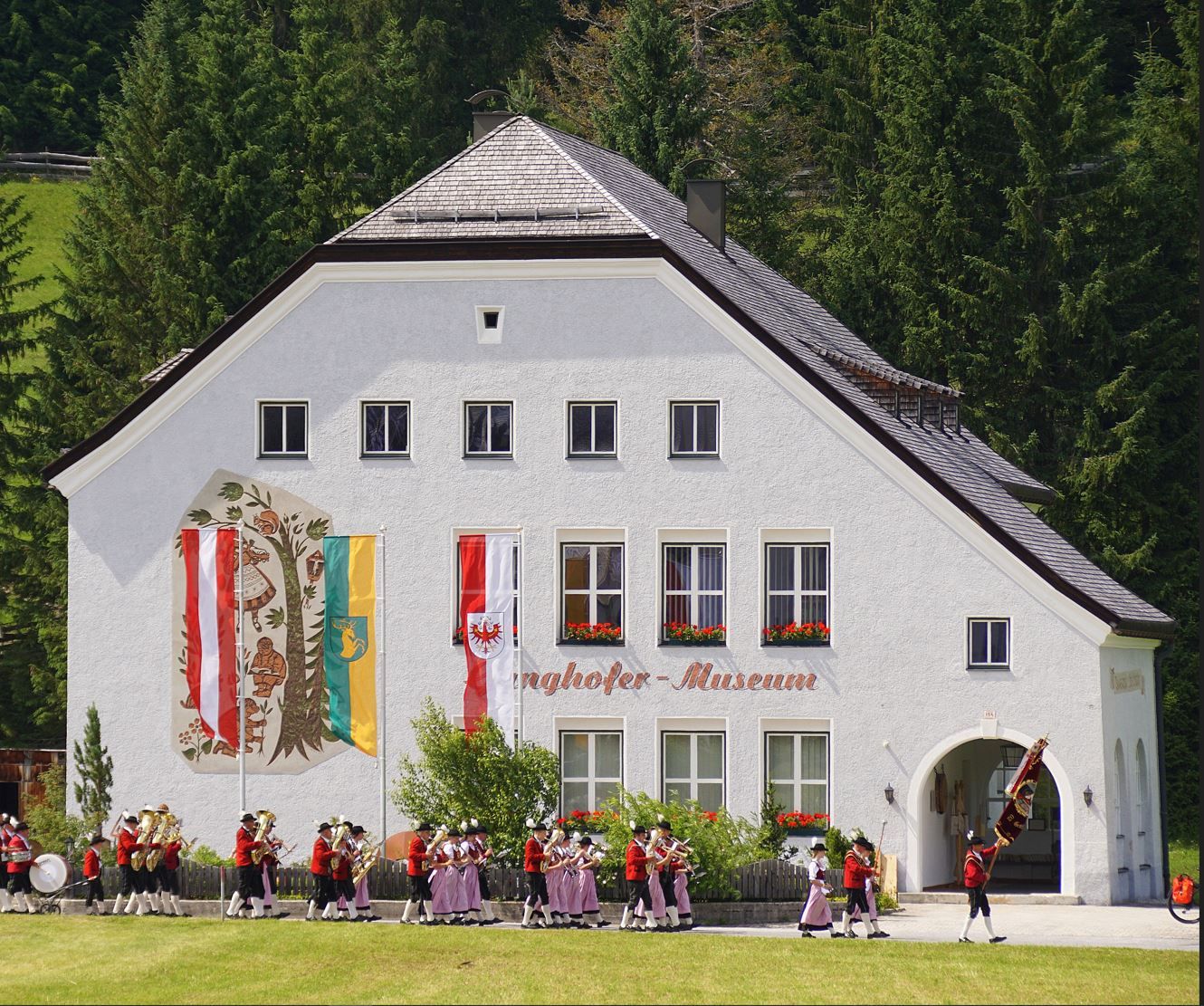 In der Nähe des Kulturhauses Leutasch steht ein weißes Gebäude mit der Aufschrift "Ponghofer Museum" und mehreren Fahnen und Wandmalereien an der Fassade, während vor dem Haus ein Spielmannszug in traditioneller Kleidung auftritt.