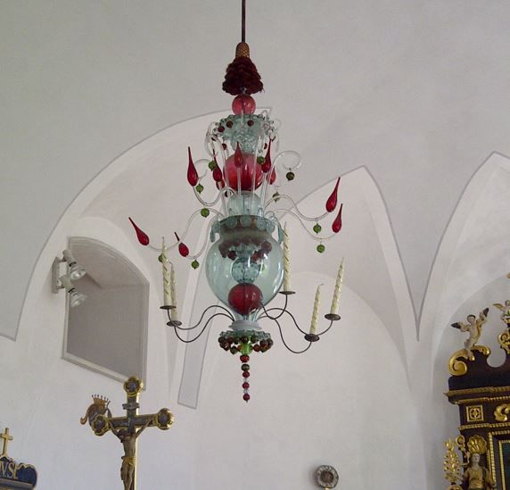 Von der weißen Gewölbedecke hängt ein verschnörkelter Glaskronleuchter mit roten und grünen Zierelementen und kerzenförmigen Lichtern herab.