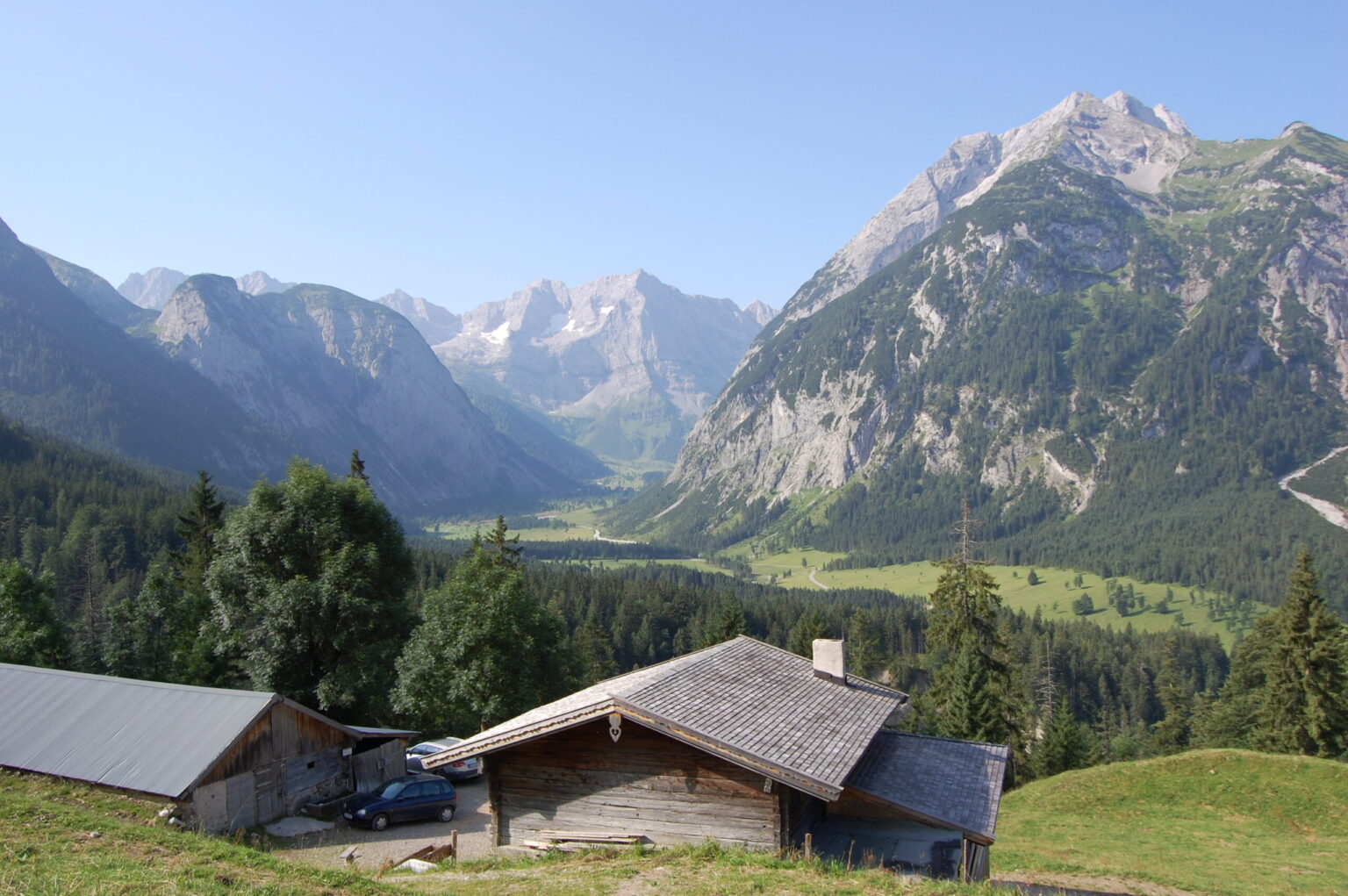 Eine hölzerne Hütte und ein Schuppen stehen auf einem grasbewachsenen Hügel mit Blick auf ein grünes Tal, dem Ausgangspunkt für die malerische Hasentalalm-Wanderung, umgeben von hohen, felsigen Bergen unter einem klaren blauen Himmel.