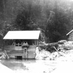 Eine kleine Holzhütte mit Satteldach steht über einem Wasserkanal am Rande eines Waldgebiets; rechts davon ist eine zweite Hütte zu sehen.