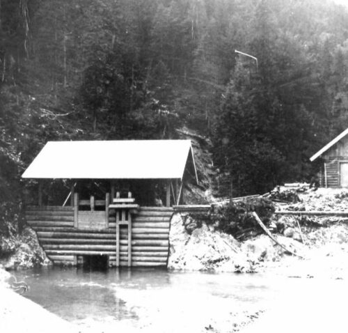 Eine kleine Holzhütte mit Satteldach steht über einem Wasserkanal am Rande eines Waldgebiets; rechts davon ist eine zweite Hütte zu sehen.