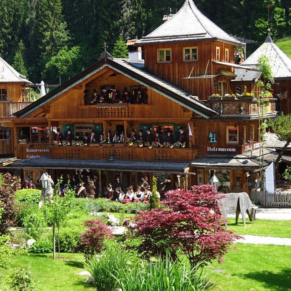 Ein großes Holzgebäude mit Balkonen und dekorativen Figuren, das als Tiroler Holzmuseum und Kunstgalerie bezeichnet wird, ist von Grünflächen und Landschaftsgärten umgeben.