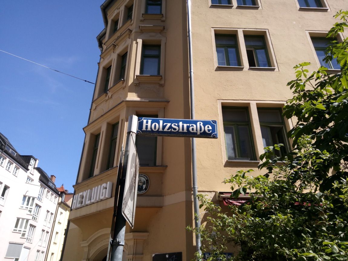 Straßenschild mit der Aufschrift "Holzstraße", ein Beispiel für einen deutschen Ortsnamen, das an einem sonnigen Tag an einem Mast vor einem beigen Gebäude mit mehreren Fenstern angebracht ist.