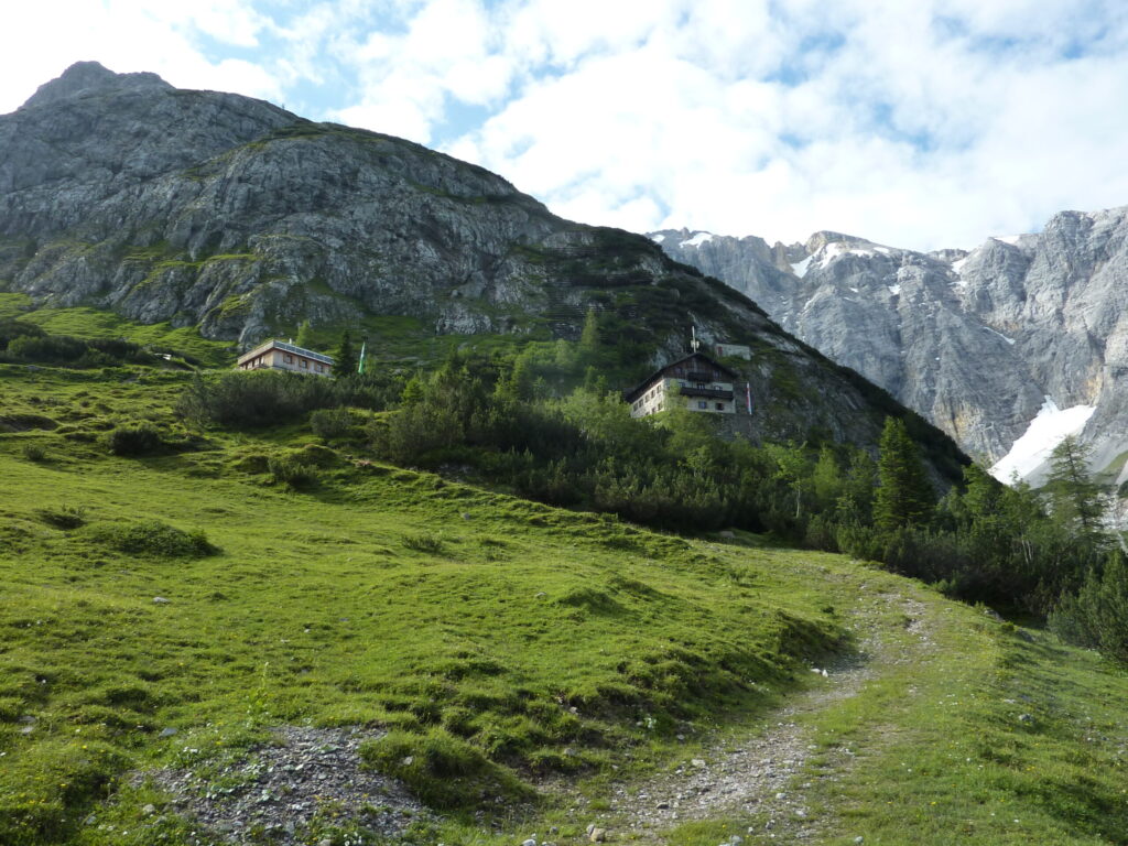 Ein grasbewachsener Berghang mit einem unbefestigten Weg, der zu zwei alpinen Gebäuden führt, darunter das Karwendelhaus, vor felsigen Hügeln und einem teilweise bewölkten Himmel.