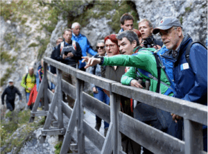 Eine Gruppe von Personen steht während einer Klimawandelexkursion Karwendelschlucht auf einer Holzbrücke in einer felsigen Umgebung. Eine Person in Grün zeigt nach vorne, während die anderen aufmerksam beobachten und die Auswirkungen des Klimawandels in den Alpen erforschen.