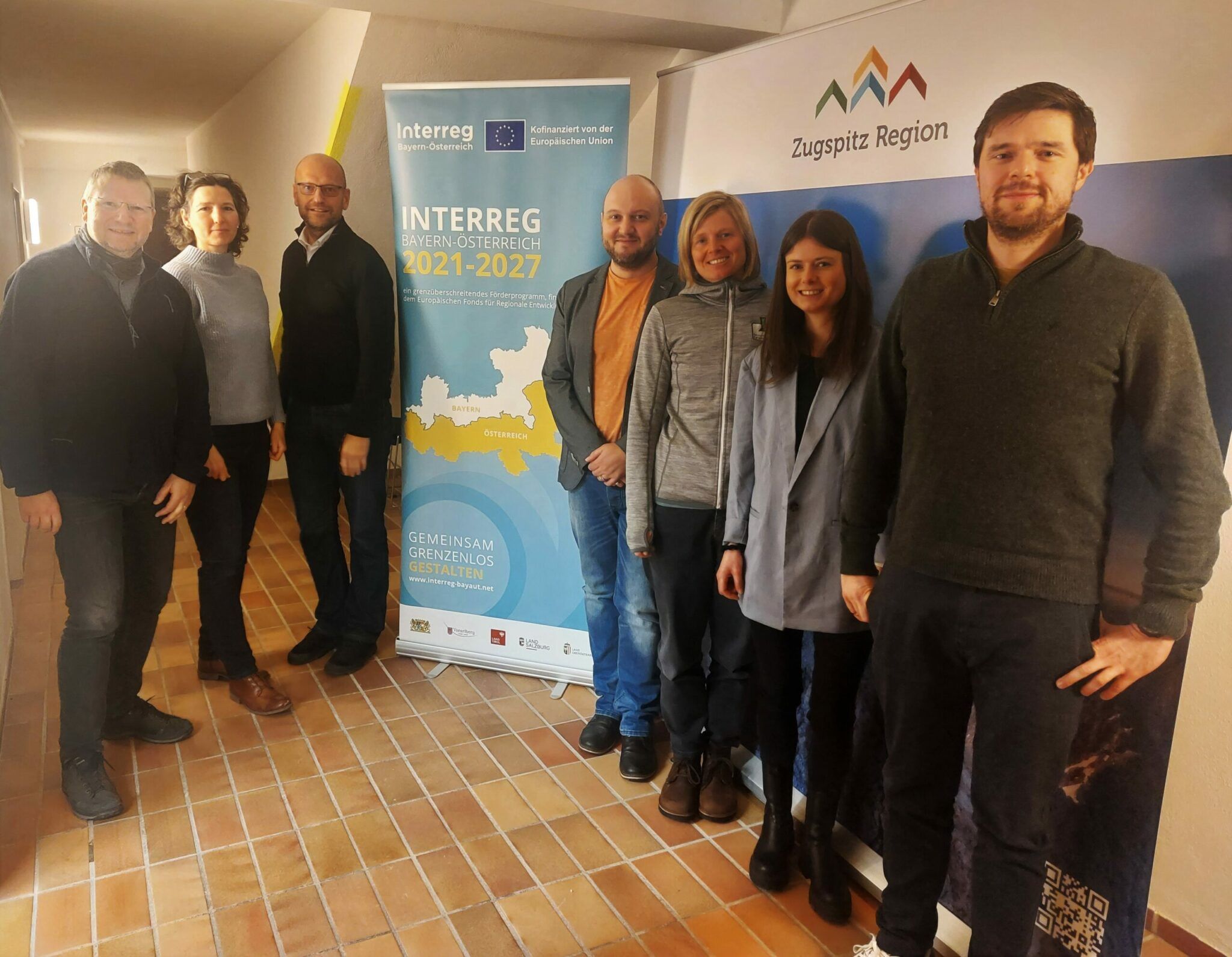 Sieben Personen stehen drinnen vor zwei Bannern für Interreg und die Zugspitzregion und posieren für ein Gruppenfoto in einem Flur mit Fliesenboden während eines Interregprojekts zum grenzenlosen Besuchermonitoring.