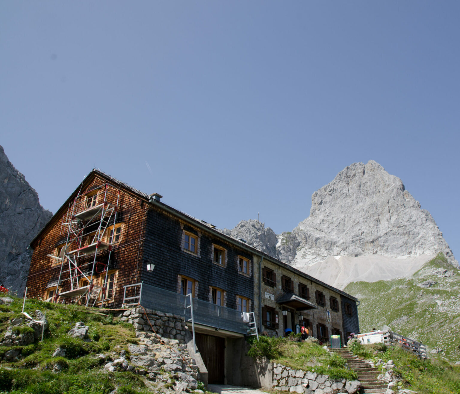 Am Fuße eines felsigen Gipfels steht unter strahlend blauem Himmel eine große, einseitig eingerüstete Holzhütte, die den perfekten Ausgangspunkt für eine Lamsenjochhütte-Wanderung darstellt.