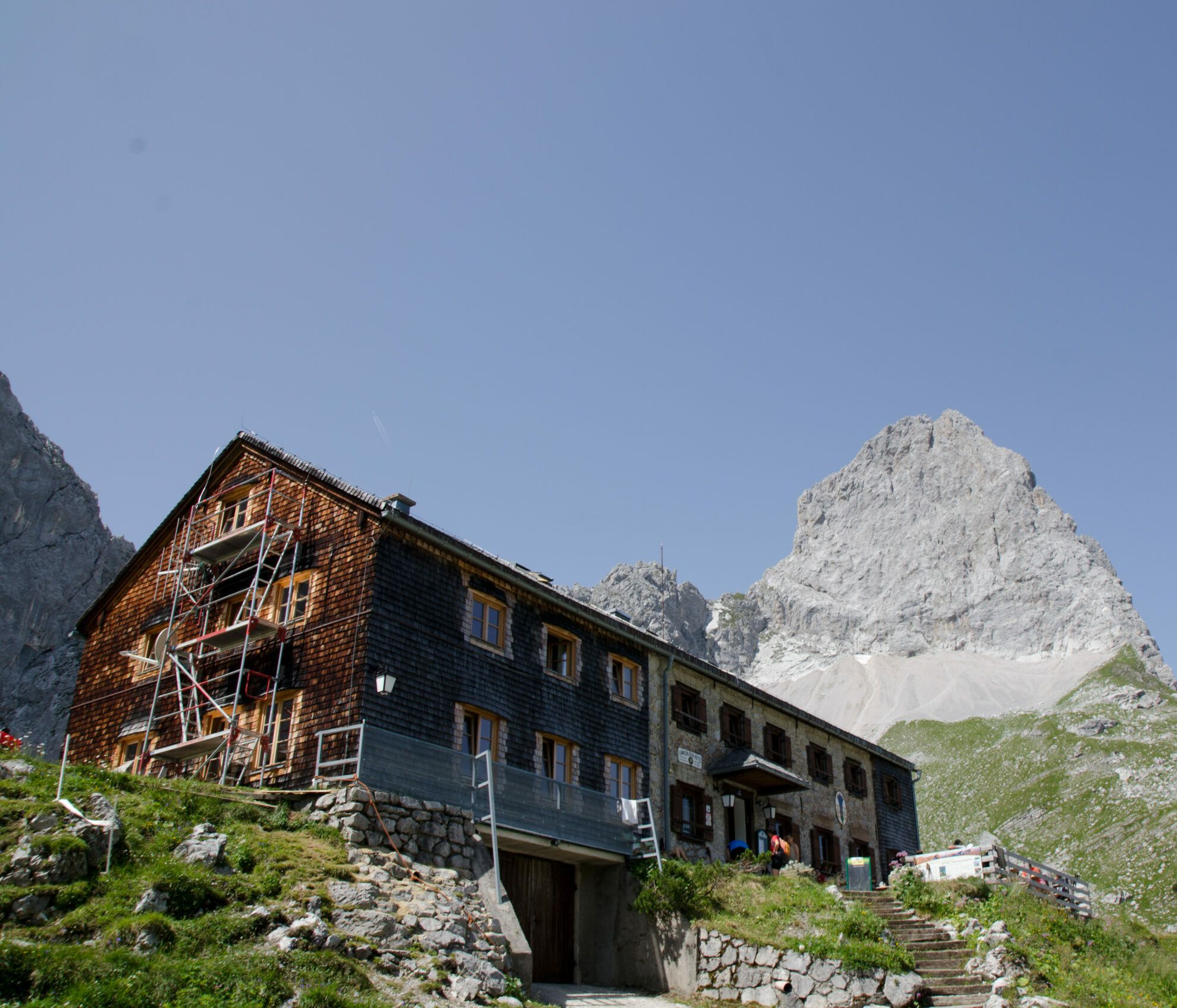 Am Fuße eines felsigen Gipfels steht unter strahlend blauem Himmel eine große, einseitig eingerüstete Holzhütte, die den perfekten Ausgangspunkt für eine Lamsenjochhütte-Wanderung darstellt.