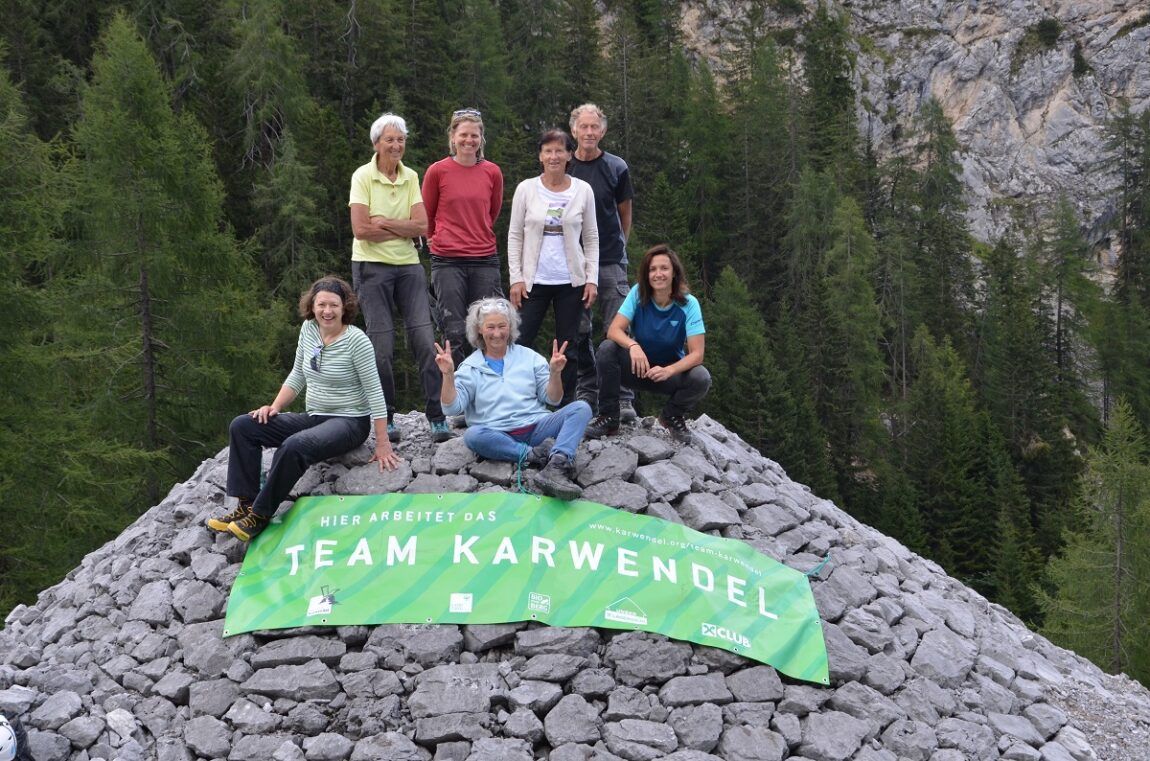 Eine Gruppe von sieben Personen posiert auf einem Steinhügel vor einem grünen Wald und dem Lawinenkegel Halltal. Vor ihnen ist ein grünes Banner mit der Aufschrift "Team Karwendel" ausgebreitet.