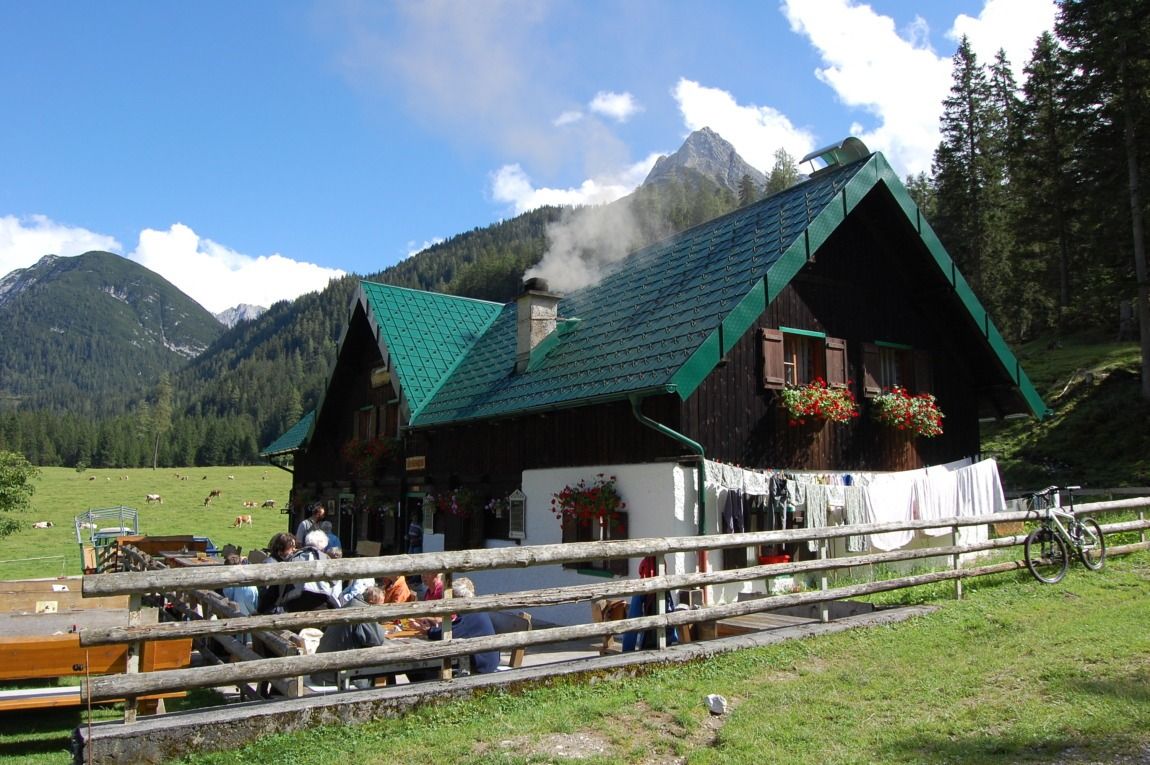 Ein Holzhaus mit grünem Dach steht auf einer Bergwiese im Alpenpark Karwendel, Wäsche trocknet auf einem Zaun und Menschen sitzen an Tischen im Freien unter freiem Himmel.
