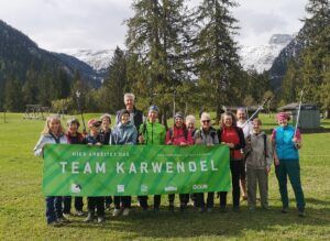 Eine Gruppe von Personen steht im Freien auf einer Wiese, hält Wanderstöcke und ein grünes "TEAM KARWENDEL"-Banner in der Hand und nimmt an einer Müllsammelaktion in den Karwendeltälern in der Nähe des Achensees teil, mit Bäumen und Bergen im Hintergrund.