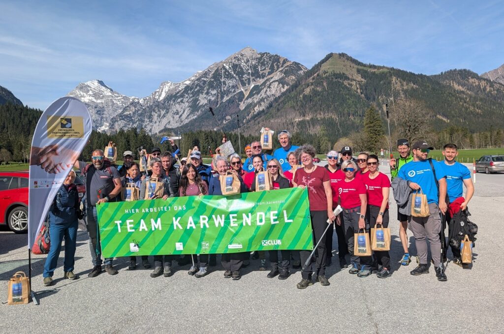 Eine Gruppe von Menschen posiert im Freien mit einem grünen "Team Karwendel"-Banner und Geschenktüten nach einer Müllsammelaktion in den Karwendeltälern am Achensee. Im Hintergrund sind schneebedeckte Berge und Bäume zu sehen.