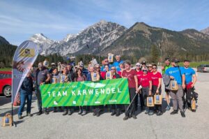 Eine Gruppe von Menschen posiert im Freien mit einem grünen "Team Karwendel"-Banner und Geschenktüten nach einer Müllsammelaktion in den Karwendeltälern am Achensee. Im Hintergrund sind schneebedeckte Berge und Bäume zu sehen.