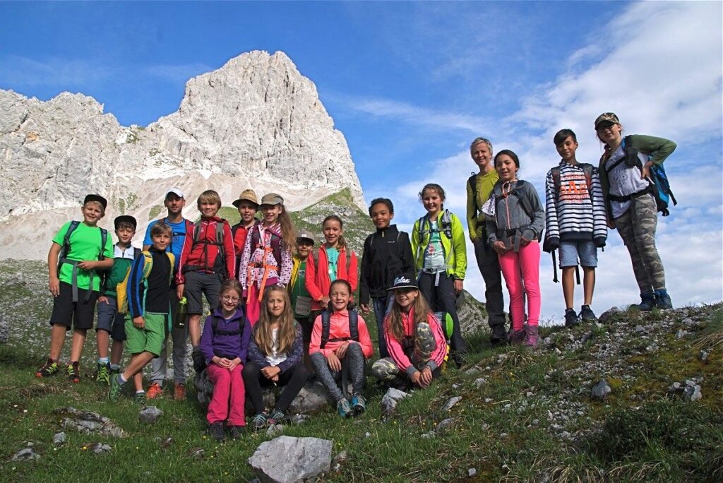Eine Gruppe von Kindern und zwei Erwachsenen mit Rucksäcken posiert für ein Foto auf einem grasbewachsenen Berghang, um das Leben im Gebirge zu erleben, mit einem felsigen Gipfel im Hintergrund unter einem blauen Himmel.