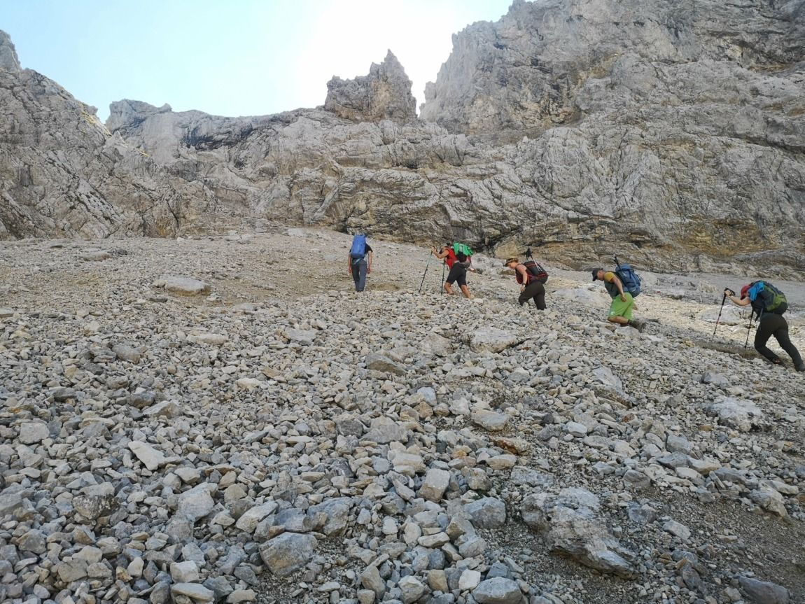 Fünf Wanderer steigen mit Rucksäcken und Trekkingstöcken einen steilen, felsigen Hang entlang des aussichtsreichen Karwendel-Höhenweges hinauf, umgeben von schroffem Berggelände und unter klarem Himmel - ein echtes Gipfeltreffen-Abenteuer.