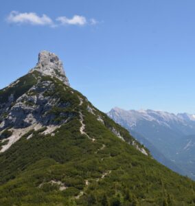 Die Arnspitzspitze erhebt sich steil und felsig über grüne, bewaldete Hänge unter einem klaren, blauen Himmel, und im Hintergrund sind ferne Berge zu sehen.