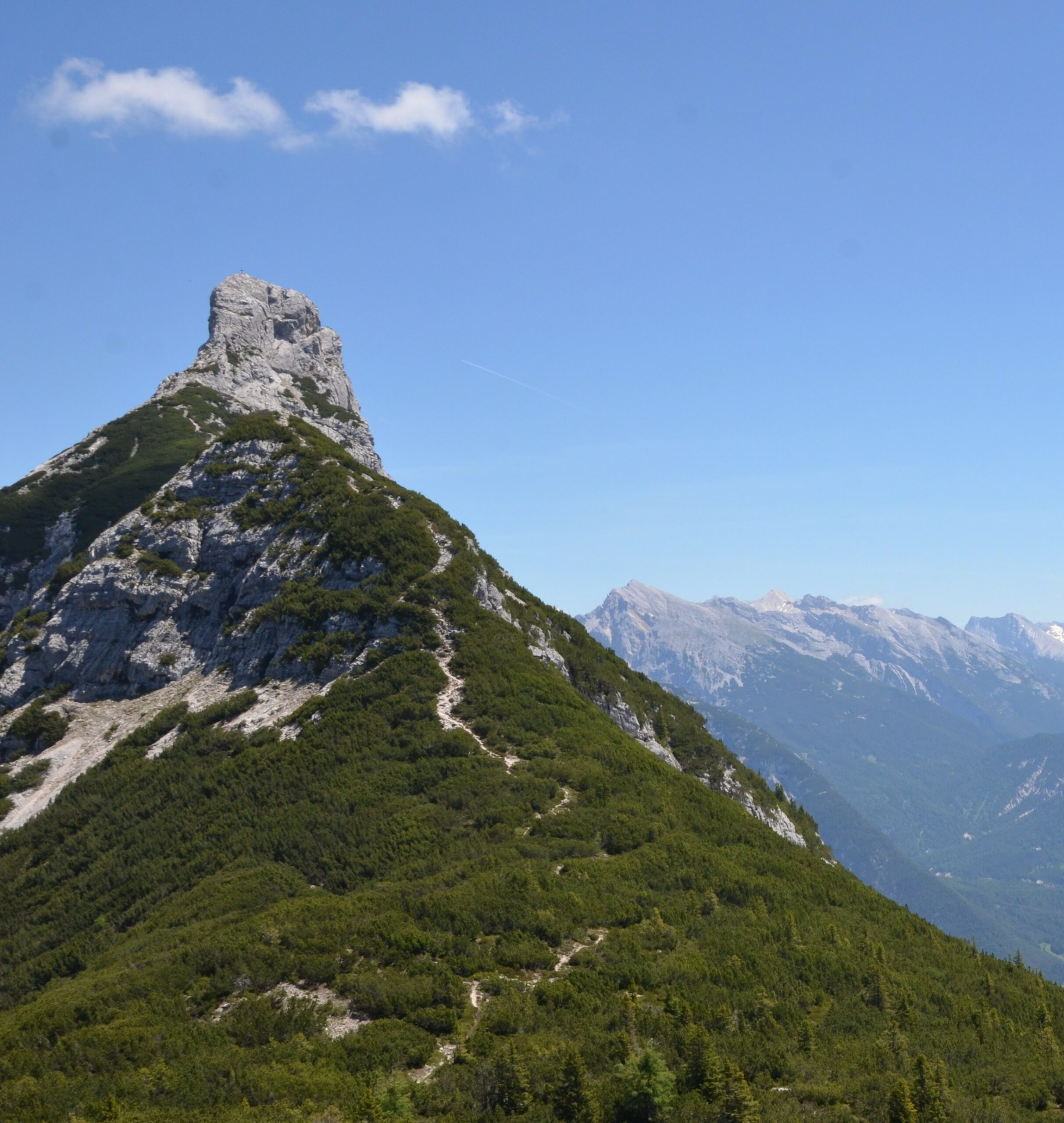 Die Arnspitzspitze erhebt sich steil und felsig über grüne, bewaldete Hänge unter einem klaren, blauen Himmel, und im Hintergrund sind ferne Berge zu sehen.