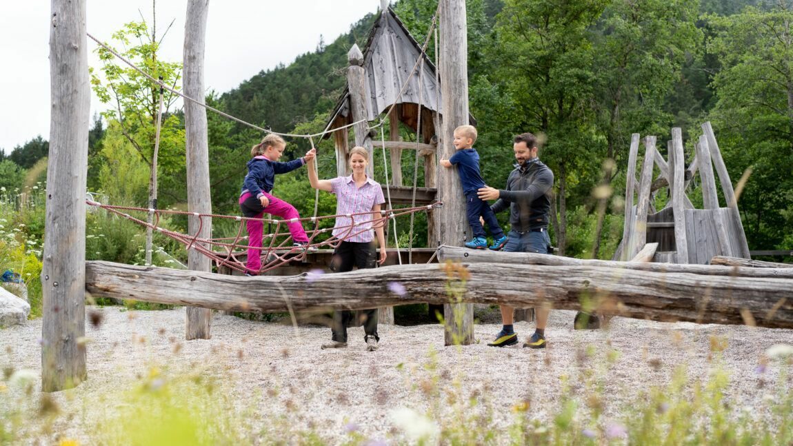 Zwei Erwachsene helfen zwei Kindern beim Spielen auf einem hölzernen Klettergerüst auf dem Spielplatz Holzerhütte, der von Bäumen und Grünflächen in der Nähe des Museums Holzerhütte umgeben ist.