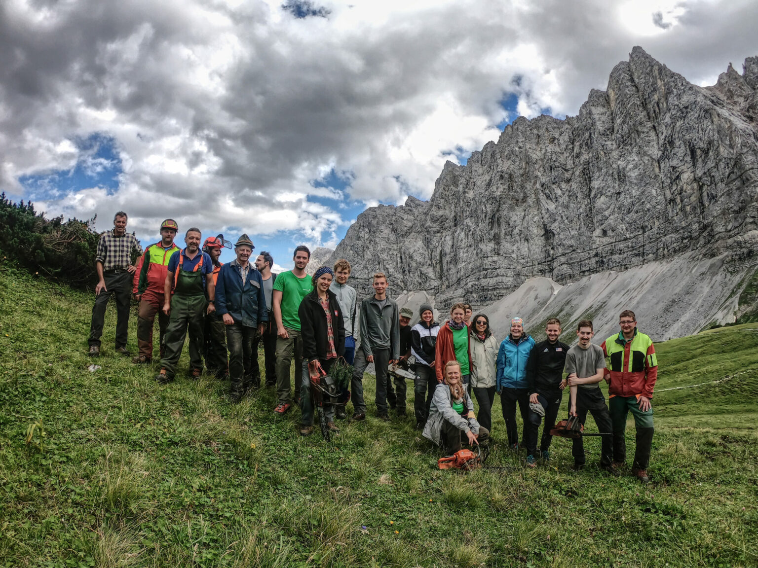 Eine Gruppe von Menschen steht mit Werkzeugen auf einem Grashang und posiert vor steilen Felsen unter einem teilweise bewölkten Himmel im Naturpark Karwendel im Rahmen der Initiative Naturschutzplan Alm.