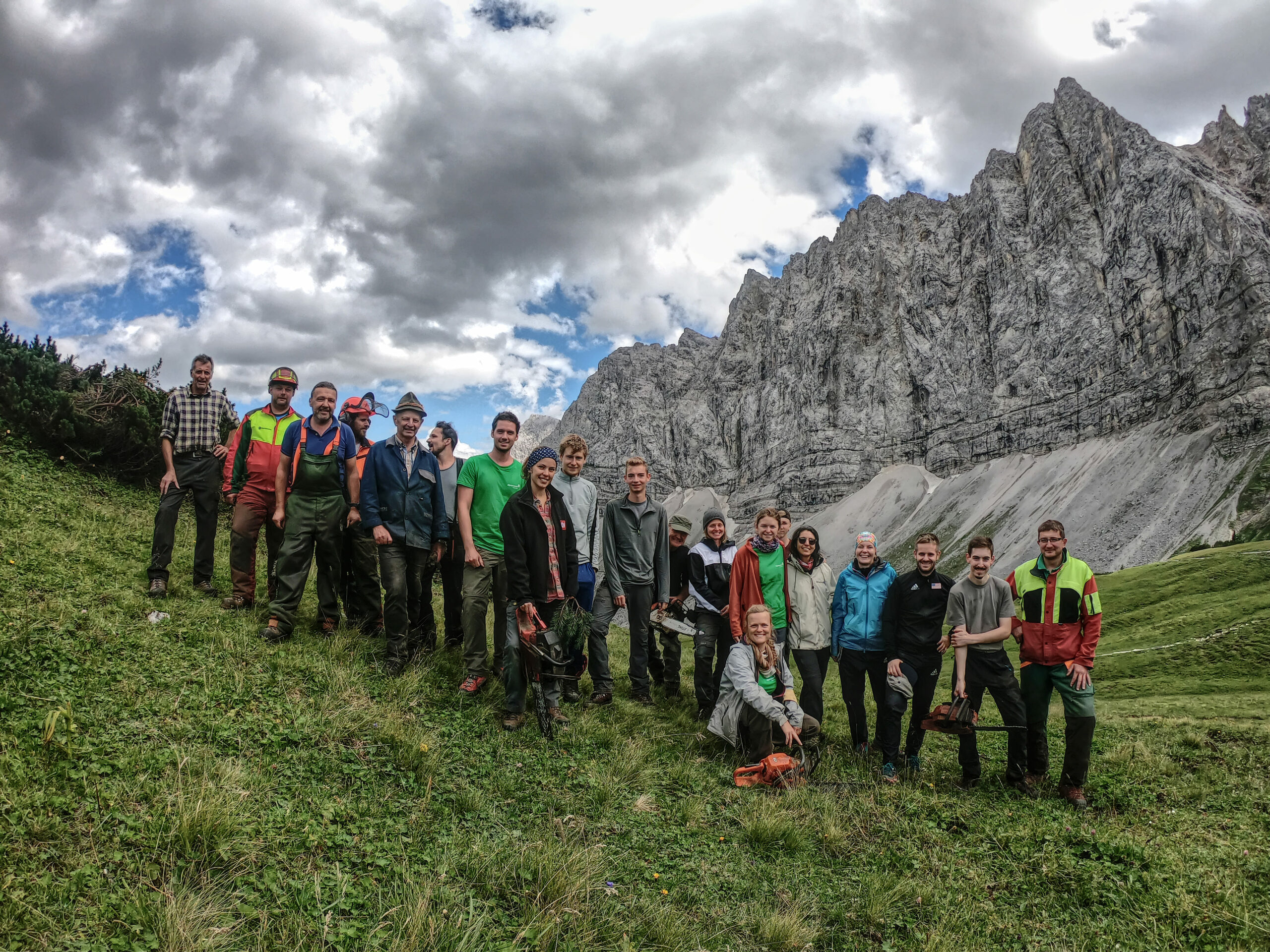 Eine Gruppe von Menschen steht mit Werkzeugen auf einem Grashang und posiert vor steilen Felsen unter einem teilweise bewölkten Himmel im Naturpark Karwendel im Rahmen der Initiative Naturschutzplan Alm.