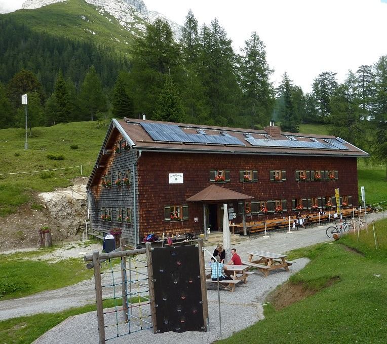 Eine Holzhütte mit Sonnenkollektoren, die der Magdeburger Hütte ähnelt, liegt in einer grünen, bergigen Gegend. Draußen versammeln sich Menschen an Picknicktischen, während davor eine kleine Kletterwand steht, umrahmt von Kiefern und Berglandschaften.
