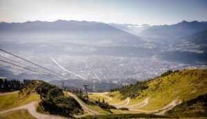 Eine kurvenreiche Bergstraße führt hinunter in ein Tal mit einem Fluss und einer Stadt, umgeben von grünen Hügeln und fernen Bergen unter einem klaren Himmel - die perfekte Aussicht nach einer lohnenden Brandjochspitze Bergtour.