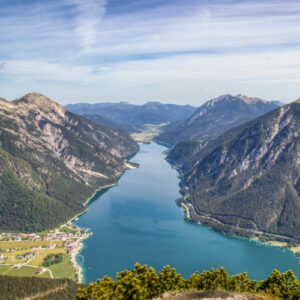 Ein breiter Fluss oder See verläuft zwischen bewaldeten Bergen unter blauem Himmel, mit kleinen Dörfern und grünen Feldern entlang des Ufers.