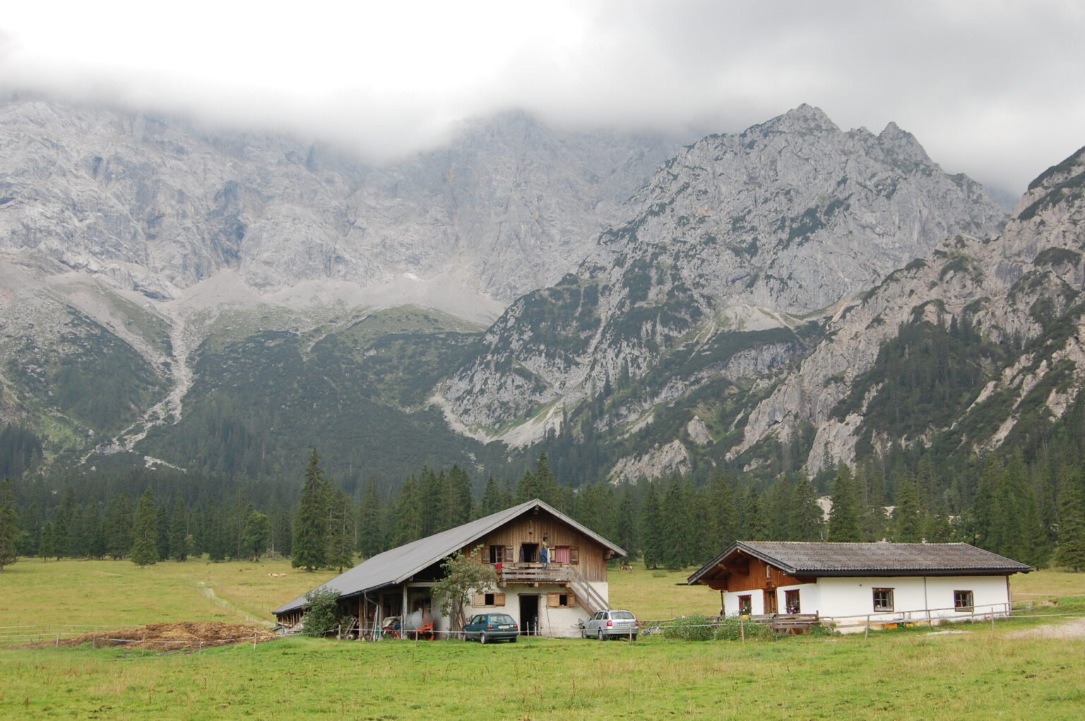 Zwei Häuser und ein Auto stehen auf einer Wiese, umgeben von Bäumen, mit hohen, felsigen Bergen und tief hängenden Wolken im Hintergrund - eine friedliche Szene auf der Rohntalalm Wanderung.