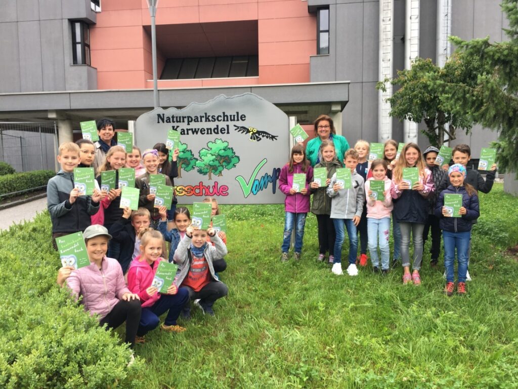 Eine Gruppe von Kindern und zwei Erwachsenen steht auf einer Wiese mit einem Kinderbuch und Broschüren in der Hand vor einem Schild der Naturparkschule Karwendel in der Nähe eines modernen Gebäudes.