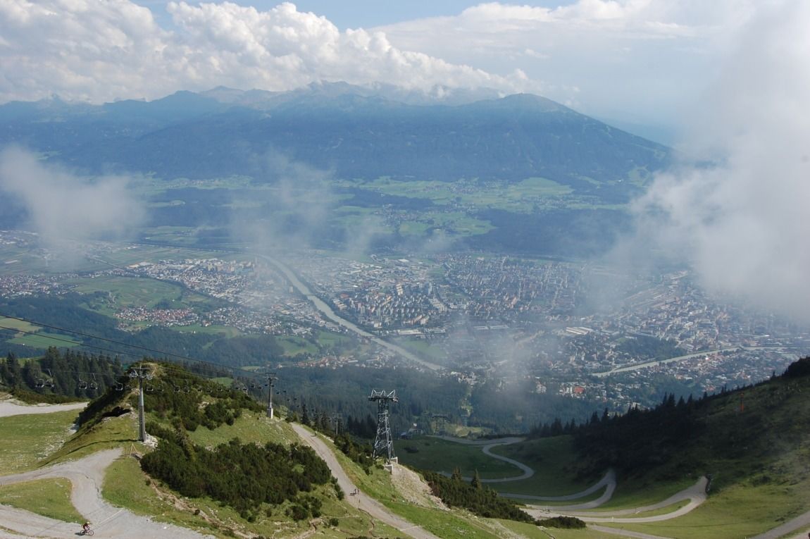 Luftaufnahme einer Stadt mit einem Fluss, eingebettet in grüne Berge und Wolken, mit kurvenreichen Straßen und der Seegrube-Seilbahn im Vordergrund.