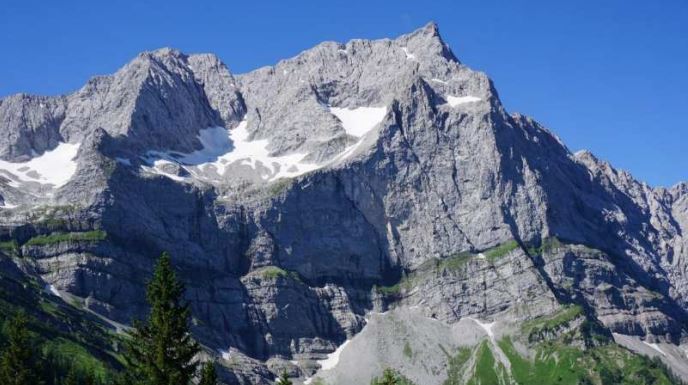 Ein hohes, felsiges Gebirge mit Schneeflecken und grünen Tannen am Fuße, unter einem klaren blauen Himmel, zeigt einen markanten Gletscher, der im Sonnenlicht glitzert.