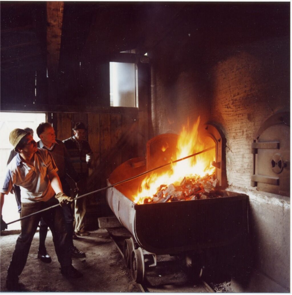 Drei Personen stehen in der Nähe eines großen Ofens, wobei eine Person das Feuer mit einem langen Werkzeug in einer industriellen Umgebung pflegt, die an Szenen erinnert, die bei einer Nature Watch Steinölwanderung beobachtet wurden.