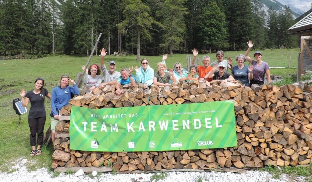 Eine Gruppe von Menschen steht hinter einem großen Brennholzstapel im Freien, hält ein grünes "Team Karwendel"-Transparent und winkt in die Kamera während ihrer Almpflege Kastenalm-Aktivitäten.