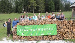 Eine Gruppe von Menschen steht hinter einem großen Brennholzstapel im Freien, hält ein grünes "Team Karwendel"-Transparent und winkt in die Kamera während ihrer Almpflege Kastenalm-Aktivitäten.