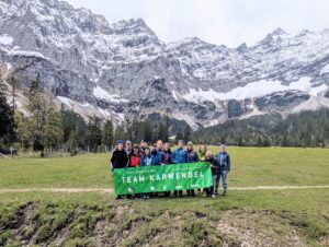 Eine Gruppe von Menschen steht im Freien vor schneebedeckten Bergen in der Nähe des Ahornbodens und hält ein grünes Banner mit der Aufschrift "Team Karwendel" in die Höhe, nachdem sie die Zaunreparaturen am Ahornboden in der atemberaubenden Alpenlandschaft abgeschlossen haben.