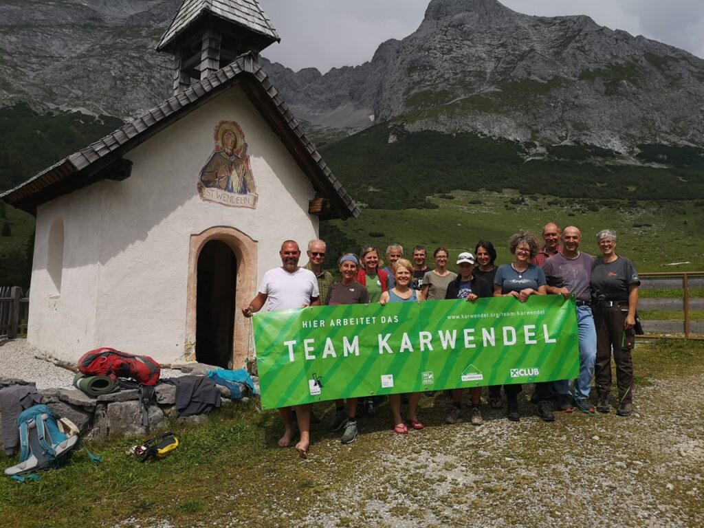 Eine Gruppe von Menschen steht vor einer kleinen Kapelle in den Bergen und hält ein grünes Banner mit der Aufschrift "Team Karwendel" in die Höhe, um ihre Bemühungen um die Almpflege Hochalm zu feiern.