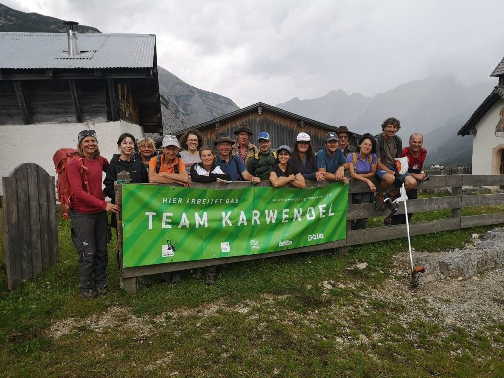 Eine Gruppe von Menschen posiert im Freien vor einem Holzzaun und Gebäuden und hält ein grünes "Team Karwendel"-Banner in die Höhe, nachdem sie die Arbeiten auf der Almpflege Hochalm abgeschlossen haben.