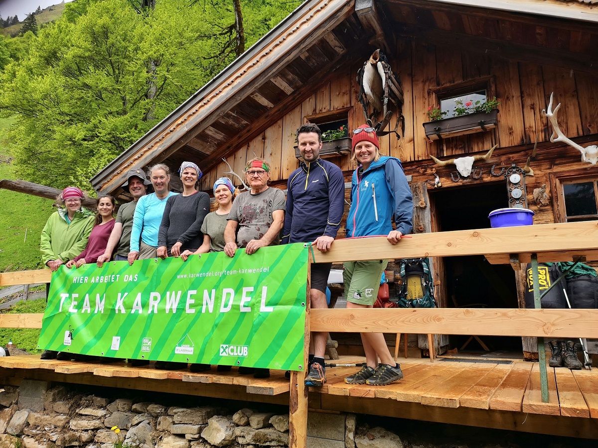 Eine Gruppe von Menschen steht und sitzt auf einer Holzveranda vor einer rustikalen Hütte und hält ein grünes "Team Karwendel"-Banner. Outdoor-Ausrüstung und Grünzeug umgeben die Hütte während ihres Almpflege Hölzelstalalm-Abenteuers.
