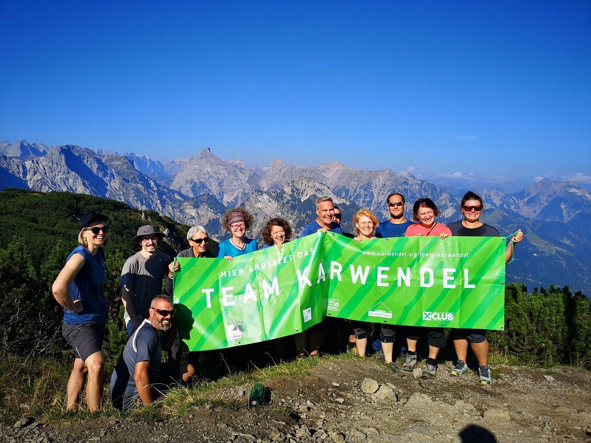 Eine Gruppe von Menschen steht auf einem Bergpfad und hält ein grünes "Team Karwendel"-Banner in die Höhe. Vor der malerischen Kulisse der Berge und des blauen Himmels unterstützen sie stolz die Almpflege Weissenbachalm.