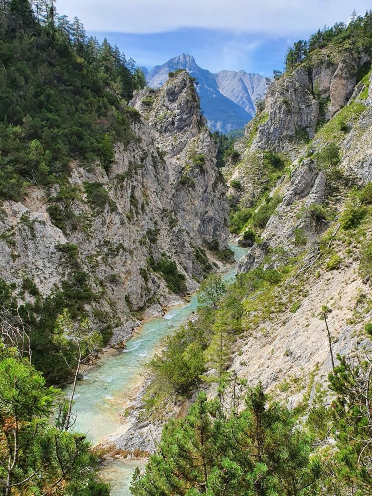 Ein schmaler türkisfarbener Fluss fließt durch die Gleirschklamm zwischen steilen, mit grünen Bäumen bewachsenen Felswänden, und im Hintergrund sind hohe Berge unter einem teilweise bewölkten Himmel zu sehen.