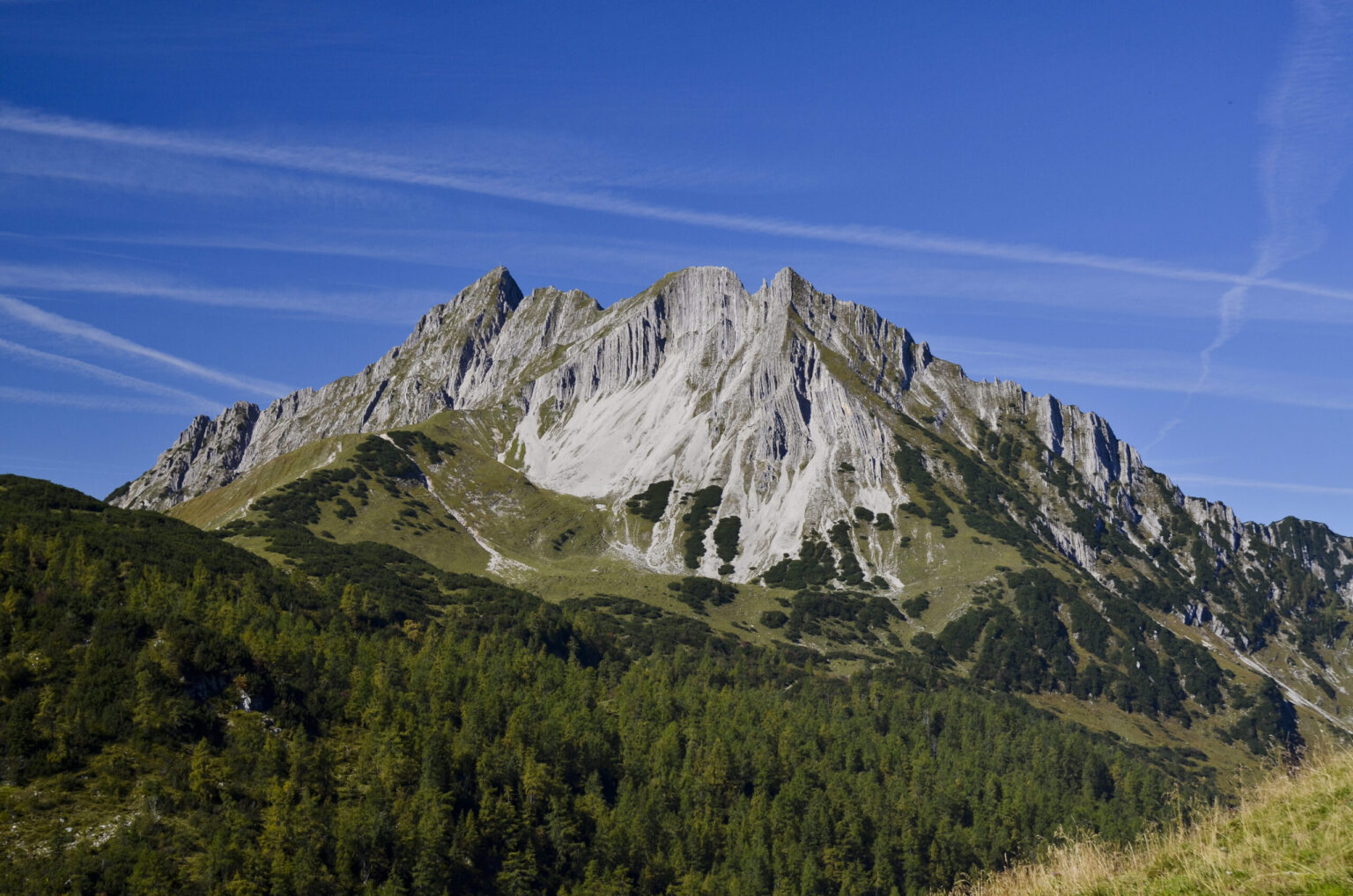 Ein schroffer Berggipfel mit steilen Felshängen erhebt sich über einem bewaldeten Gebiet unter einem klaren blauen Himmel und lädt Abenteurer ein, die malerische Mondscheinspitze Wanderung zwischen dünnen Wolkenschlieren zu erleben.