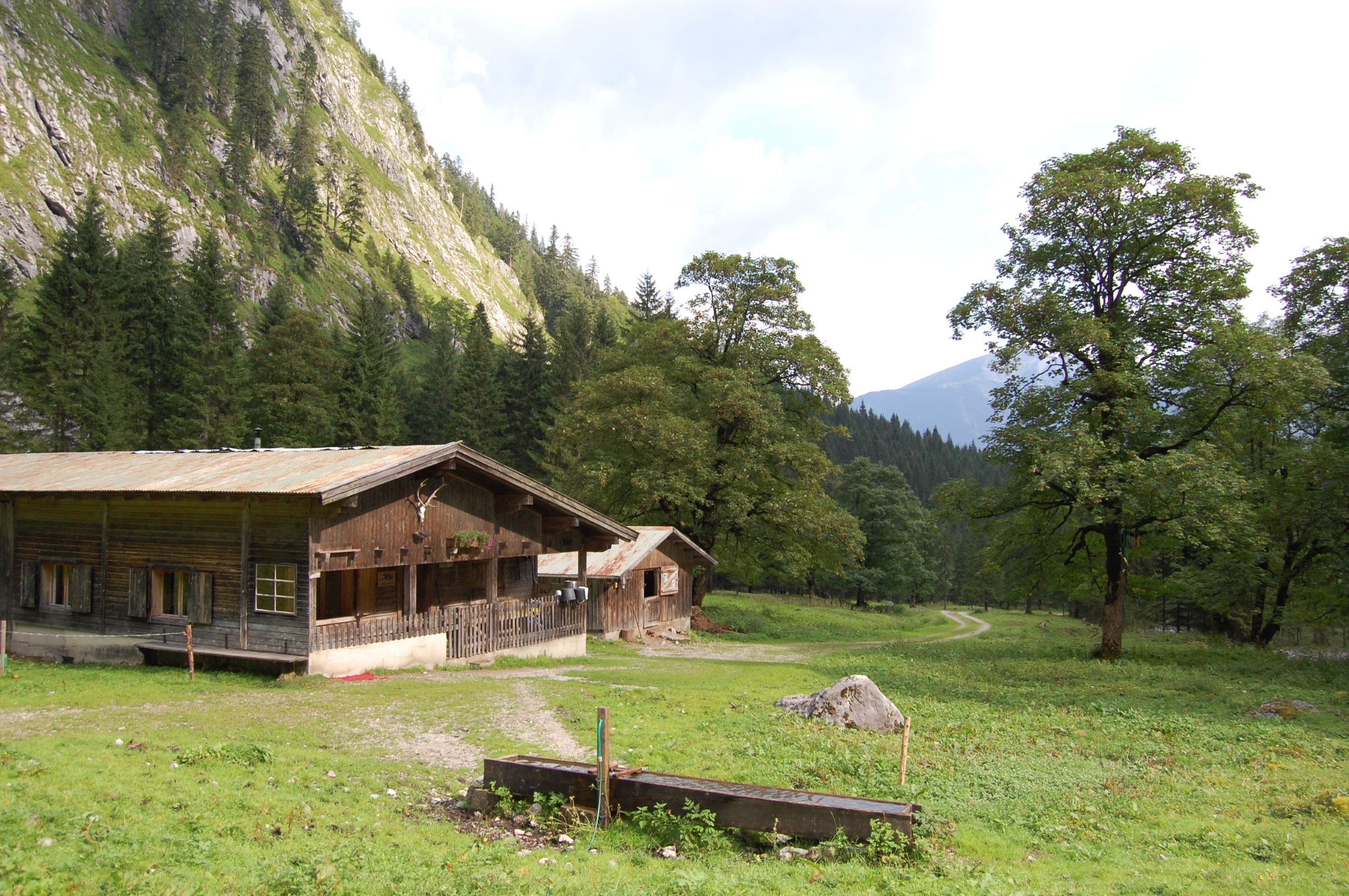 Eine Holzhütte mit schrägem Dach steht auf einer Wiese in der Nähe eines bewaldeten Berges und lädt die Besucher ein, ihre Tortalwanderung entlang der Bäume und des in die Ferne führenden Feldwegs zu beginnen.
