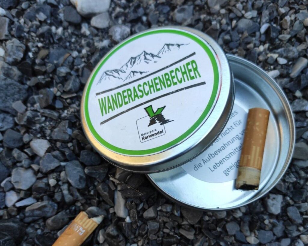 Ein kleiner Metallbehälter mit der Aufschrift "WANDERASCHENBECHER" steht offen auf dem Kies und enthält zwei benutzte Zigarettenstummel, die vielleicht von Sportschützen zurückgelassen wurden, die eine Pause vom Schützentraining machten.